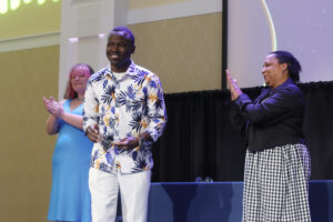 A photo of a smiling student on stage receiving an award, next to a dean and another student who are both clapping.