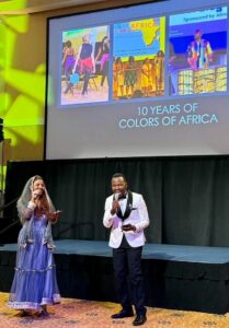 A student in a tuxedo holding a microphone and another student in traditional dress on stage, with a screen behind them that reads '10 Years of Colors of Africa.'