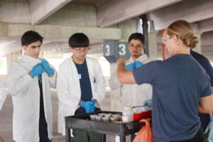 Hannah Steele '26, who earned a Noyce Scholarship at UMW, demonstrates to high schoolers Zubair Mohammadzai (left), Michael Young (center), and Maiwand Barakzai (right) how to soak the cotton balls in washable paint before throwing them at black canvases hanging in UMW's Alvey Parking Deck. Photo by K Pearlman Photography.