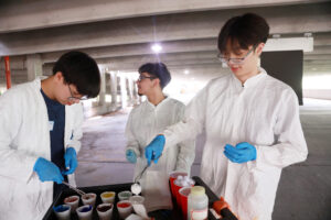 From left: High schoolers Michael Young, Alexander Rosas, and Vincent Phan dip cotton balls in washable paints before lobbing them at black canvases to create artwork resembling evolving stellar clusters, which they learned about in an astrophysics lesson. Photo by K Pearlman Photography.