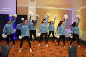 Members of the UMW Pep Dance group, wearing light blue UMW sweatshirts, black tights, and sneakers, perform a cheer with white pom-poms.