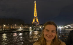 Carly Radgowski, a senior French and environmental science major at the University of Mary Washington, stands in front of the Eiffel Tower at night. Smiling college-aged woman with blonde hair in the bottom right corner of the screen with the Eiffel Tower lit up in the background.