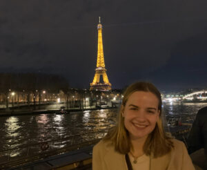 Carly Radgowski, a senior French and environmental science major at the University of Mary Washington, stands in front of the Eiffel Tower at night. Smiling college student with medium-length blonde hair in the bottom right corner of the screen with the Eiffel Tower lit up in the background.