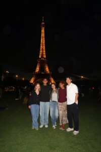 Description: Carly Radgowski (left) with a group of four friends from the MICEFA program, in front of the Eiffel Tower, which is lit up at night. Caption: Carly and her friends she made while studying abroad visiting the Eiffel Tower at night. Photo courtesy of Carly Radgowski.