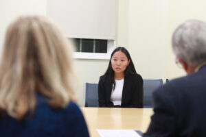 A college student in a suit answers a question of two judges with their backs to the camera.