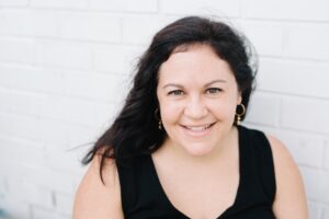Jessica McCaughey '01 smiling against a white brick background. 