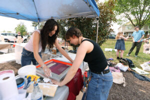 A student helps another student print a shirt under the tent.