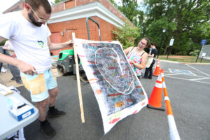 A professor and student look at the student's print on cloth with the steamroller in the background and orange plastic cones in the foreground.