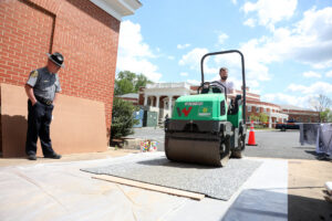 A professor rides a steamroller over a print and woodblock covered in foam, while UMW's police chief watches.