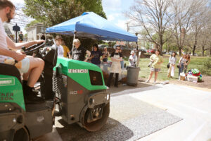 A professor rides the steamroller while students watch from a small tent and a line next to the tent.
