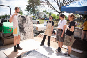 A professor holds up a print to show three students with a steamroller in the background.