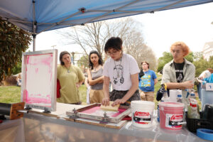 Students watch as one screen-prints a T-shirt under a tent.