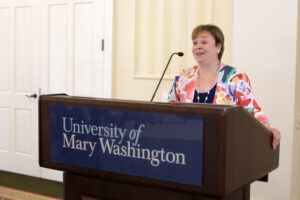An alumna talks into a microphone at a podium with a University of Mary Washington sign on it.