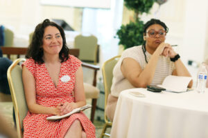 Two alumni sit at a table listening to presentations. One holds a small book and a pen.