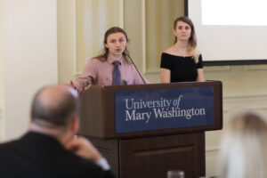 A student speaks into a microphone at a podium with a University of Mary Washington sign on it. Another student stands to the student's left side.