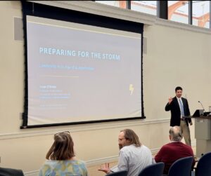 An alum speaks in front of a screen that reads: Preparing for the Storm: Leadership in an Age of AI Acceleration. Sean O'Brien, AI Expert-in-Residence, University of Mary Washington. Several UMW faculty and staff with their back to the camera in the foreground. 