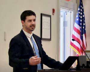 An alum in a suit and tie speaks to an audience with an American flag in the background. 
