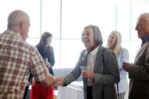 An alumna, holding a cup in one hand, shakes hands while talking to several professors at a reception. Her husband is next to her.