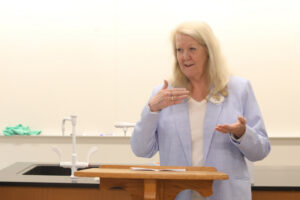 A smiling professor stands at the front of the room, talking and gesturing with her hands.