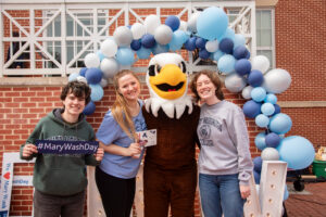 Three students stand under a blue, white, and gray balloon arch with UMW mascot Sammy D. Eagle on the Cedric Rucker University Center patio.