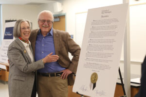A couple standing with their arms around each other, next to the Board of Visitors Resolution on a giant poster.