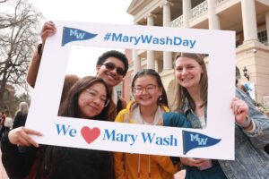 A group of four students in front of the Cedric Rucker University Center holding up a frame that reads #MaryWashDay and We love Mary Wash.