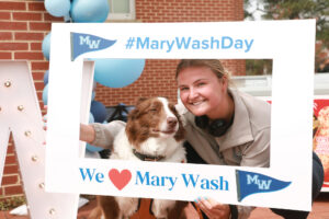 A student and her dog pose for a picture with a sign that says #MaryWashDay and We love Mary Wash.