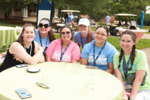 Six young alumnae in T-shirts sitting at a table outside of Monroe Hall.