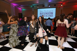 Alumni dancing at the all-class party on a black-and-white checkerboard floor with a screen that says 'Mary Washington Reunion Weekend.'