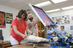A printmaking professors teaches an alumna how to use screenprinting equipment in an art studio.