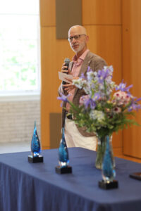 UMW President Troy Paino holding a microphone in Seacobeck Hall's Weatherly Wing. In front of him is a table with flowers and several alumni awards on it.
