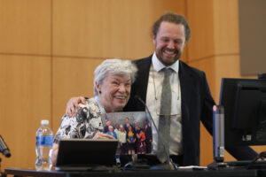 A 1962 alumna thanks her alma mater for a UMW alumni award and is joined by a 2015 alumnus whom she worked with. She holds a photo of herself former Virginia Governor Glenn Youngkin and other public officials at the time the legislation passed.