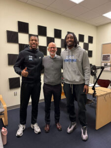Two UMW men's basketball players smiling for the camera with President Paino in the center in the HCC podcast studio.