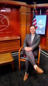 A UMW student in a suit and glasses, seated in a chair in the U.S. Department of State with a red background and an American flag behind her.