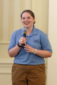A student wearing a blue polo shirt and black pants, holding a microphone and smiling.