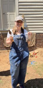 A photo of a student wearing overalls, a T-shirt, and a ballcap holding a trowel and a fragment of a ceramic plate.