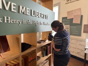 A student in a blue striped sweater labeling objects in a museum. The exhibit reads: "Give me liberty! Patrick Henry in the Fight for Freedom."