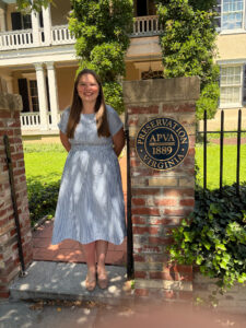 A student wearing a summery dress, smiling and standing next to a brick column with a round plaque next to an iron fence. A historic building is in the background.