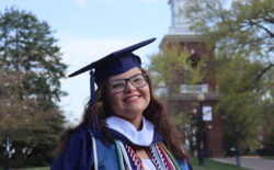Student wearing a graduation cap and gown and glasses, smiling in front of the UMW Bell Tower.
