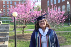 A student in a blue cap and gown, smiling and standing on the UMW campus with brick buildings and pink blossoms in the background. 
