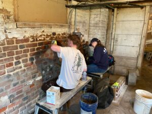 Two students sitting on a bench with their backs to the camera, applying mortar in the basement of the Mary Washington House.