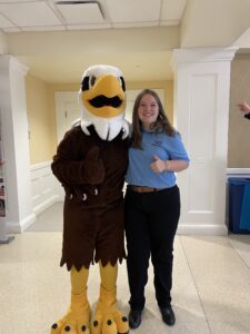 A photo of a student wearing a blue polo shirt and black pants smiling and holding her thumb up with her arm around UMW mascot Sammy D. Eagle.