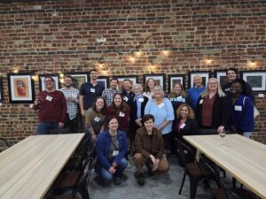 A large group of people standing against a brick wall in a winery. Paintings are on the wall, lights are overhead, and two tables flank the group.