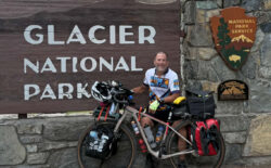 A photo of a man and his bicycle in front of the Glacier National Park sign with evergreen trees in the background.
