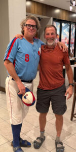 A man in an old Montreal Expos baseball uniform, holding a baseball cap, with his arm around a man in a red T-shirt, black shorts, and sandals.