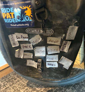 A saddlebag covered in duct tape stickers with names written on them. A Ride Pat Ride sticker is in the top left corner.