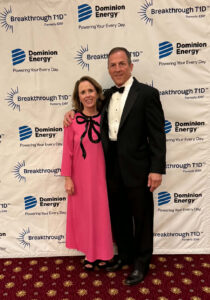 A man in a tuxedo and a woman in a pink dress with a black bow at the top, standing in front of a step-and-repeat at a gala for Breakthrough TD1.