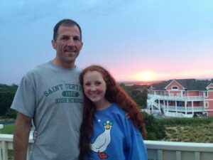 A man and his daughter standing on a balcony in front of a sunset with a coral-colored beach house in the right background.