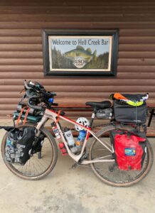 A photo of a packed bicycle in front of a log cabin-style wall with a sign that reads Welcome to Hell Creek Bar.