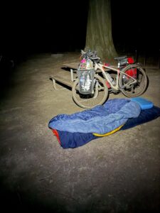 A photo taken at night of a bicycle with a sleeping bag rolled out in the dirt in front of a picnic table and a tree.
